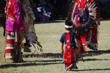 A small child in traditional Native American attire walks among adults who are also dressed in traditional Native American attire