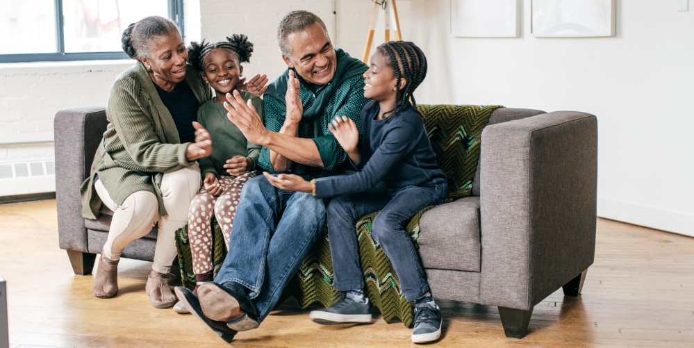 A Black grandmother, grandfather, elementary-age girl, and elementary-age boy sit on a couch and members of the family look at each other and give each other high-fives
