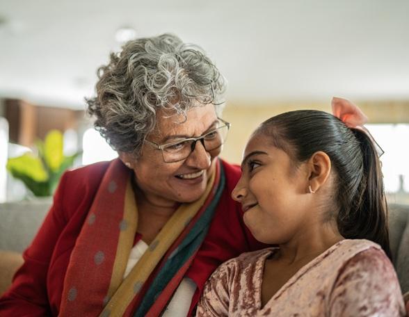 A Mexican grandmother and granddaughter smile as they look at each other and lean into each other while sitting on the couch together