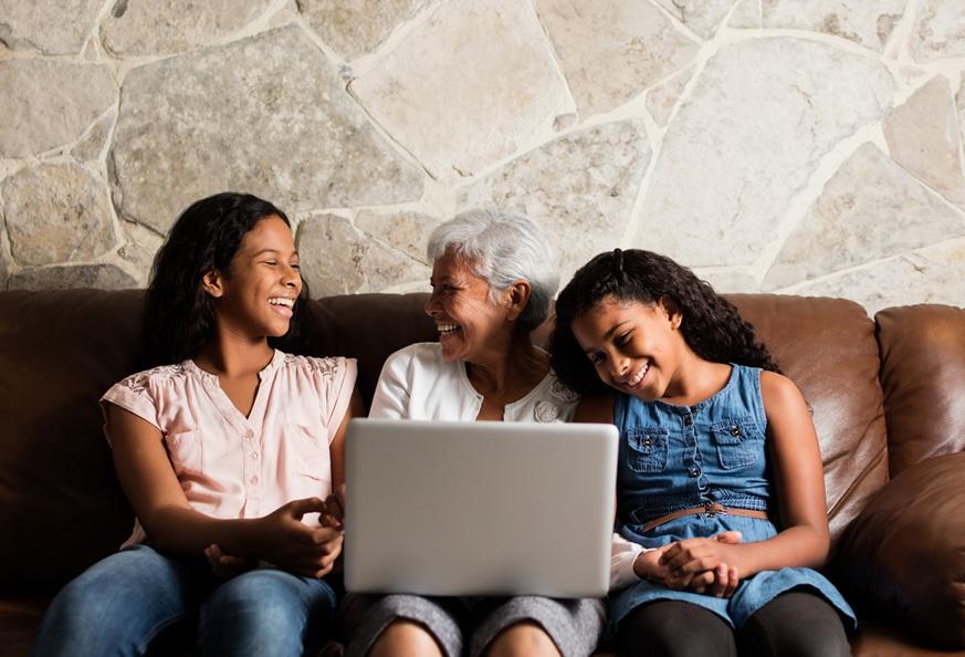 A Latina grandmother and her two tween and teen granddaughters smile as they sit on a couch together with a laptop