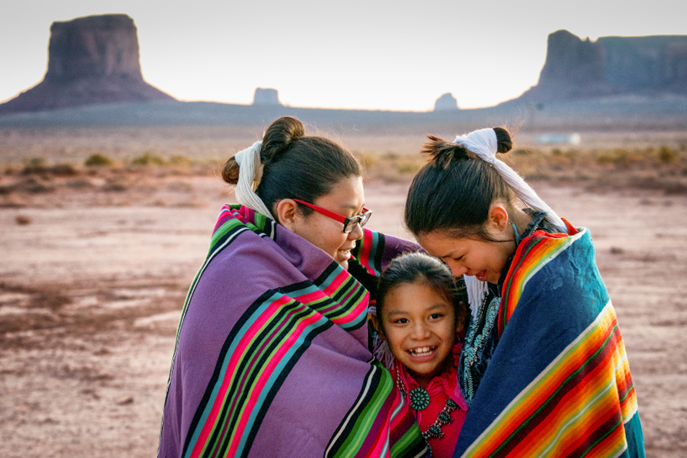 In Monument Valley, two older Navajo sisters face inward and look at their younger sister, who is in between them. All three are smiling and in traditional dress, with handwoven traditional blankets around their shoulders.