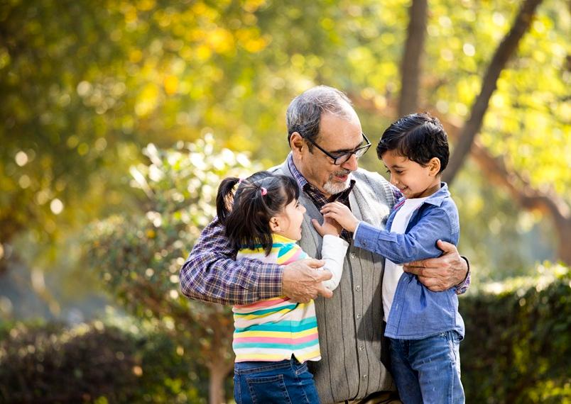 In a park, an Indian grandfather kneels to embrace his young granddaughter and grandson, who are both smiling and placing their hands on his heart
