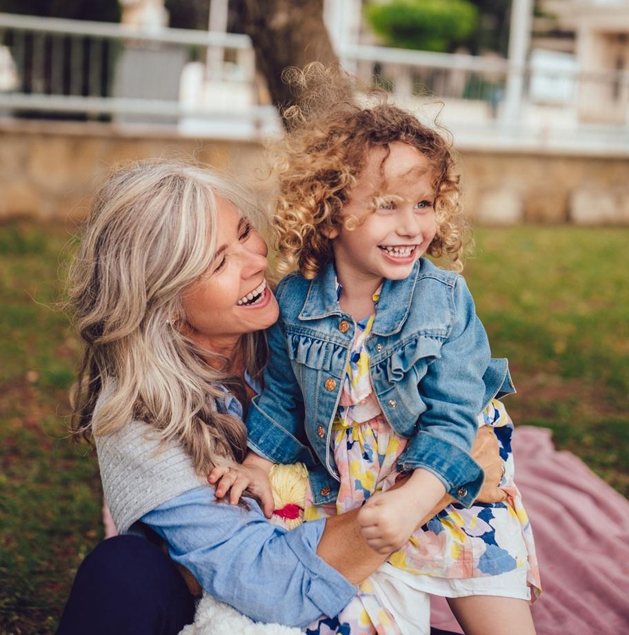 A white grandmother and her young granddaughter smile as the grandmother sits on the ground and hugs her standing granddaughter