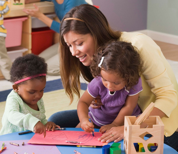 A young child care worker smiles as she watches two toddler girls draw together. One child is in her lap and the other is standing next to the table. 