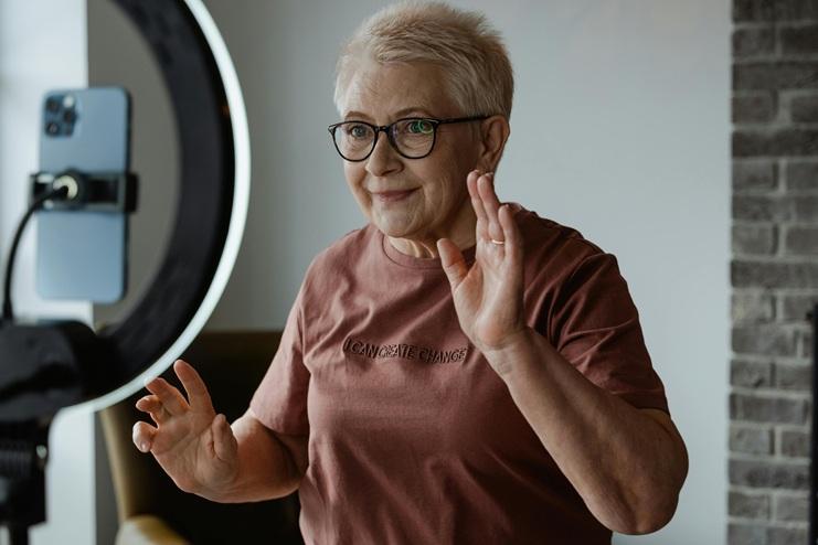 An older woman gestures as she films herself using a smartphone and a ring light