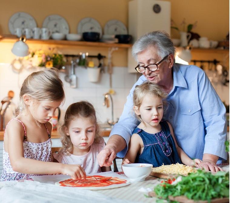 A white grandmother helps her three young granddaughters make homemade pizza the kitchen