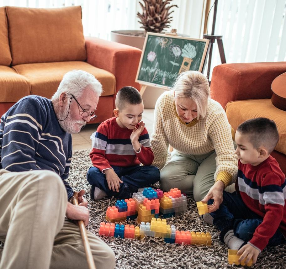 A white grandmother and grandfather sit on the floor with their twin grandsons, playing with large, Lego-type blocks