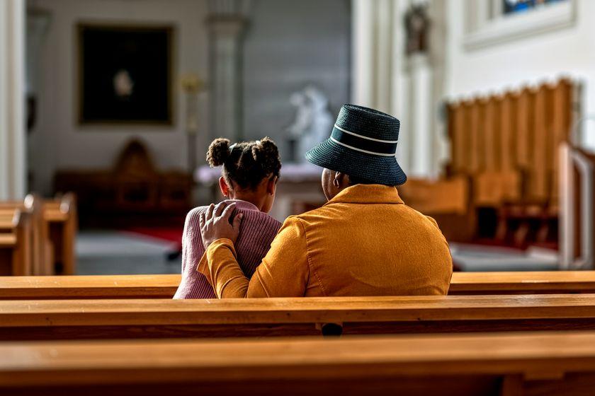 A photograph from behind of a Black grandmother and her granddaughter in a sanctuary pew