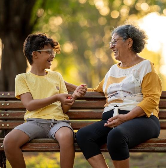A Latina grandmother and her grandson, both wearing glasses, sit on a park bench and smile as they look at and talk with each other