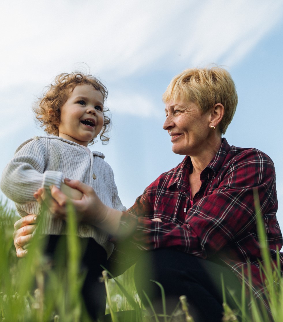 A white grandmother and her young grandchild stand in a field together. The grandmother is sitting or kneeling and looking at and holding onto her grandchild and the child is smiling.