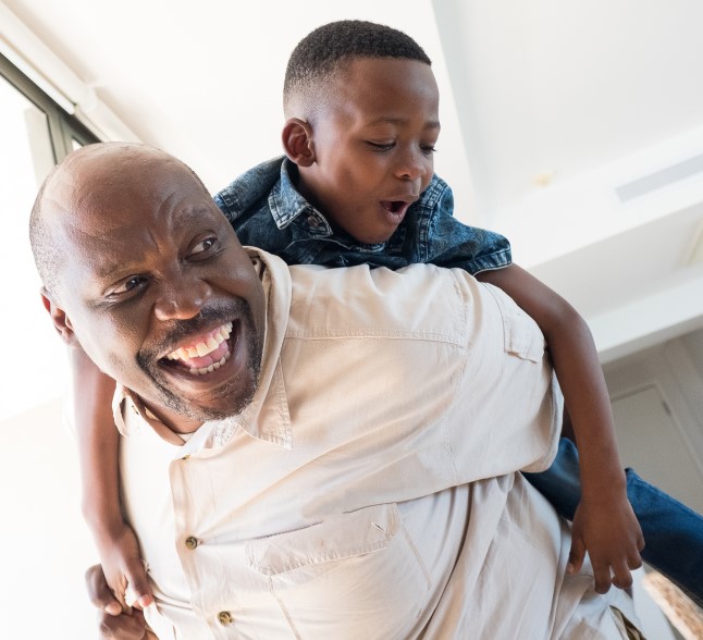 A Black grandfather smiles widely as he plays with his young grandson, holding the joyful child on his back