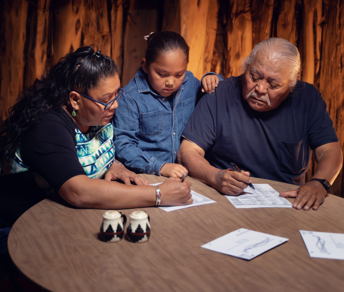 A Native child stands between two older Native adults as they sit at a table and fill out paperwork