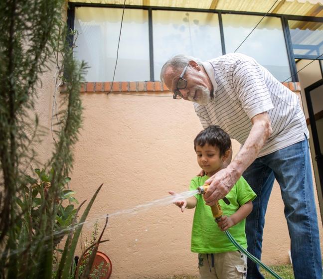 A Latino grandfather and young grandson use a garden hose to water plants together outside