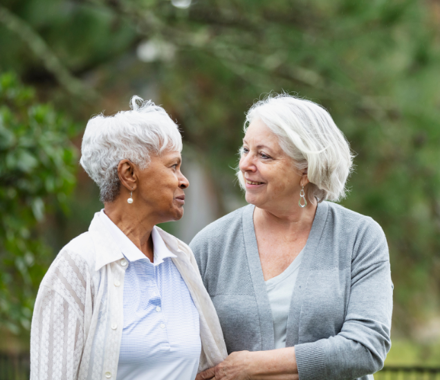 Two older women, one Black and one white, stand outside and look at each other. One woman is holding onto the other's arm in a gesture of empathy.