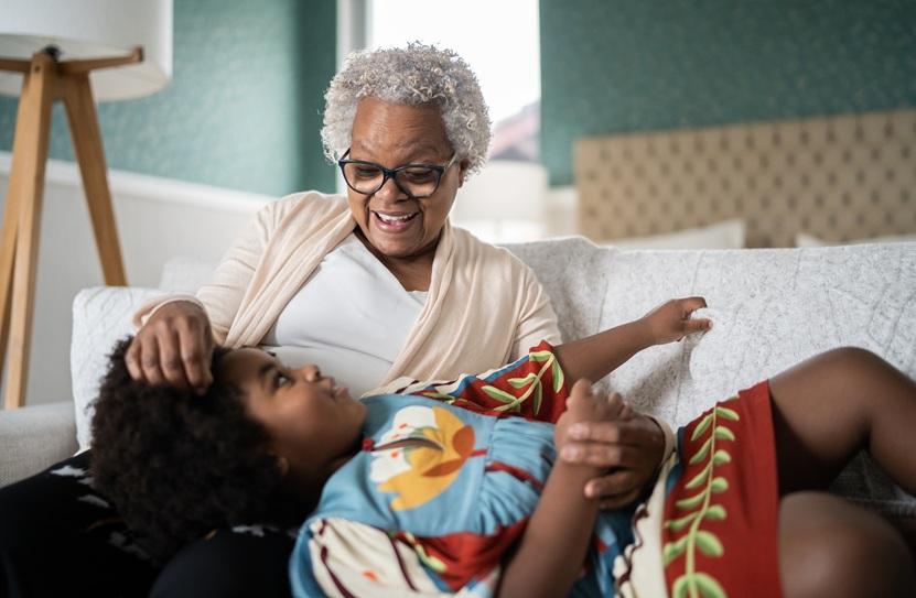 A Black grandmother smiles at her granddaughter. The granddaughter is looking up at her grandmother and has her head in her grandmother's lap