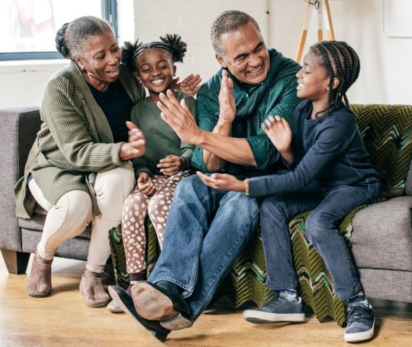 An African American grandmother and grandfather high-five their granddaughter and grandson while all four sit on a couch