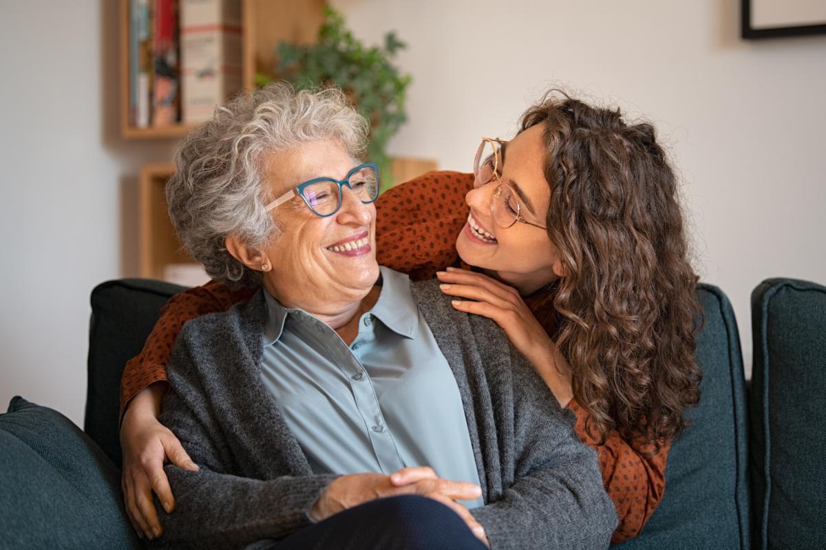 A white grandmother sits on a couch and smiles at her teenage or young adult granddaughter, who is leaning over the back of the couch, and embracing her grandmother and smiling at her.