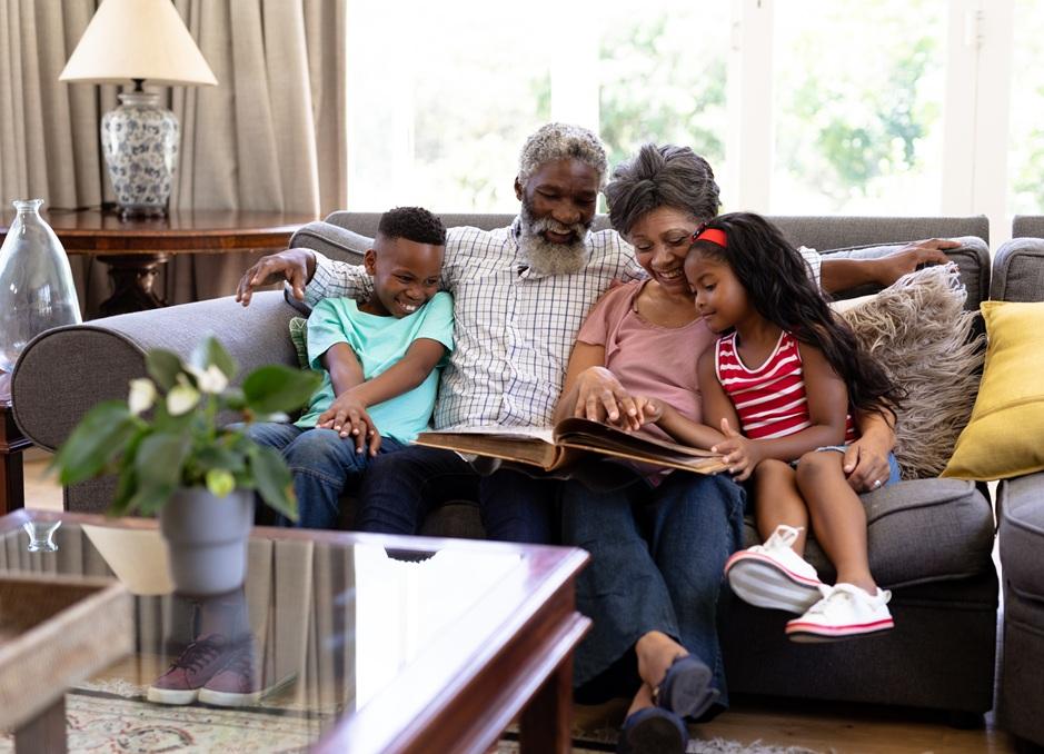 A Black grandfather and grandmother sit together on a couch and smile as they look through a book or photo album with their grandson and granddaughter