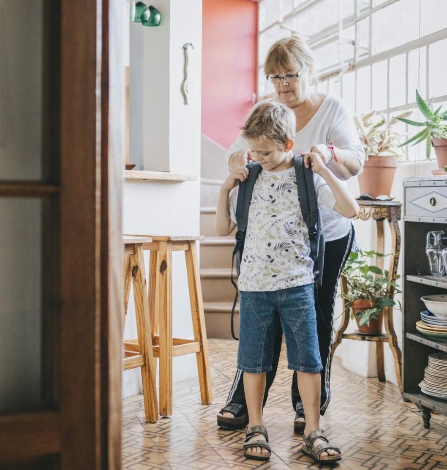 A white grandmother helps her grandson put his school backpack on