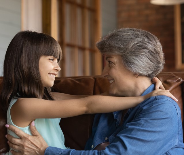 A Latina grandmother and her elementary-aged granddaughter smile at each other with their arms around each other