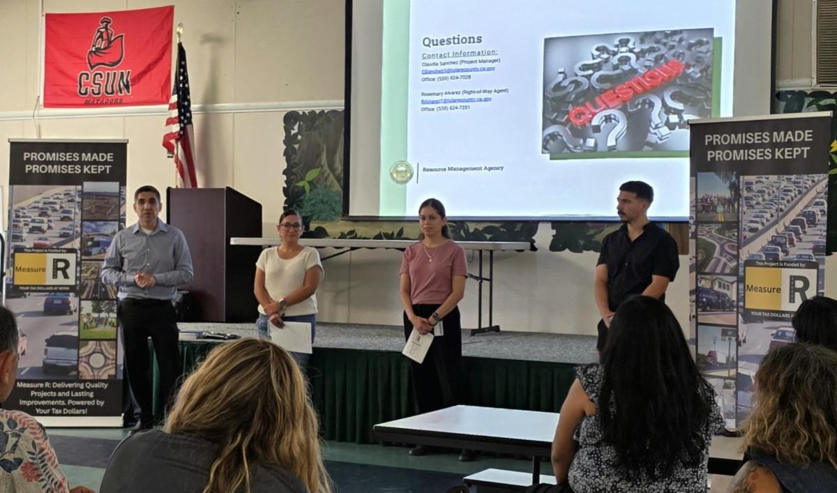 RMA staff stand at the front of a room during a community meeting at Palm Elementary School in Orosi. A presentation about the Road 130 pedestrian improvement project is displayed behind them as residents listen from their seats.