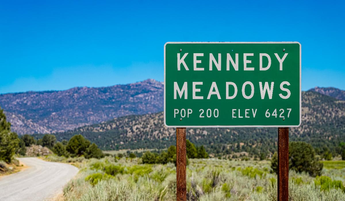 A green roadside sign welcomes visitors to Kennedy Meadows, California, listing a population of 200 and an elevation of 6,427 feet, with the Sierra Nevada mountains in the background.