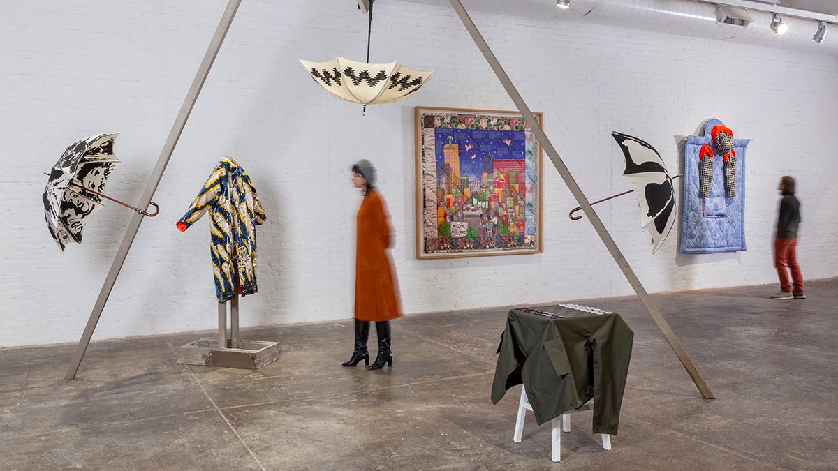 A photo of a gallery installation featuring umbrellas displayed from a large thin armature. Two free-standing garments are displayed below while two other pieces are displayed on the far wall. Two visitors are seen looking at the art.