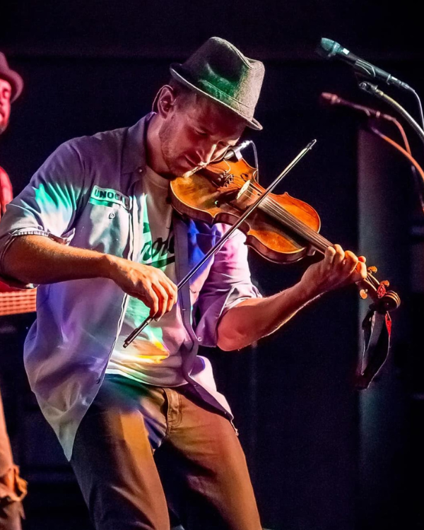 Focused musician in a fedora plays violin on a dark stage, bathed in colorful lights beside a microphone, creating an energetic mood.