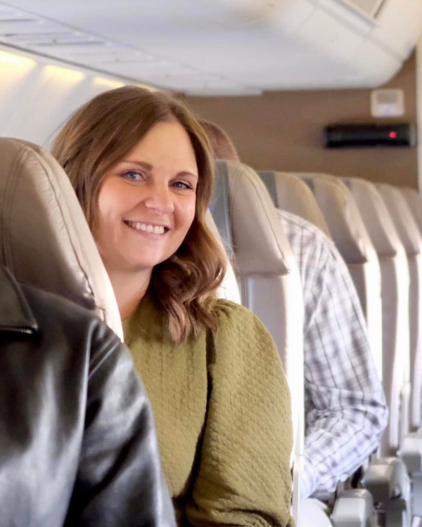 Smiling woman in an olive-green sweater sits in a commercial airplane cabin, with leather seats and another passenger behind her.