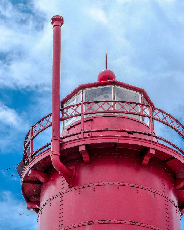 Close-up of a bright red lighthouse lantern room and chimney, with ornate railings and riveted metalwork against a cloudy blue sky.