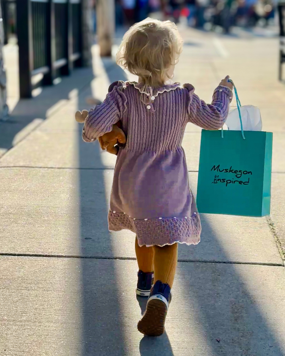 A young child walks along a sunlit sidewalk, carrying a turquoise â€œMuskegon Inspiredâ€ shopping bag and a small toy; the scene feels warm and cheerful.