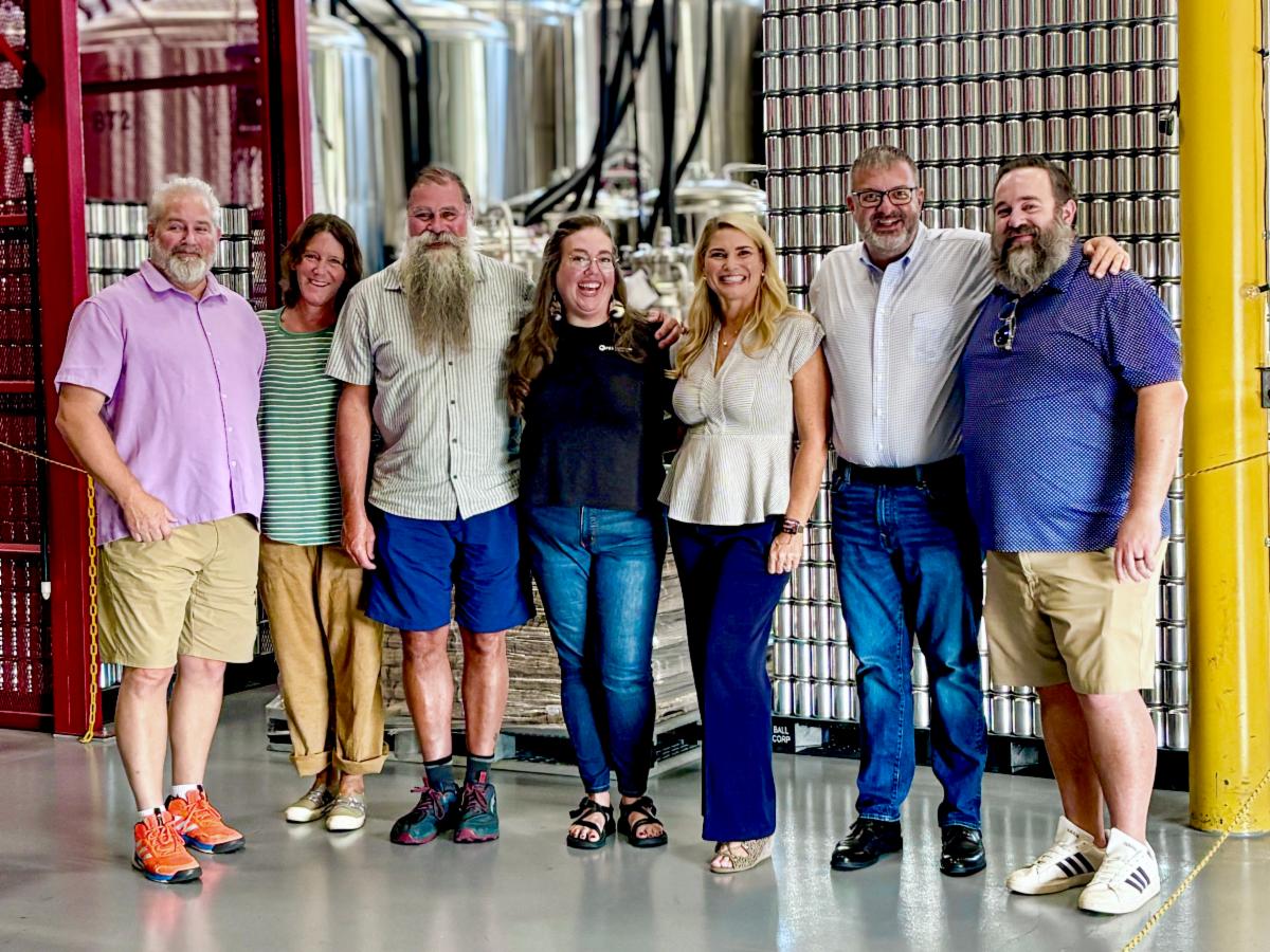 Seven smiling adults pose together in a bright industrial facility, with metal tanks and pipes behind them, conveying a friendly, welcoming mood.
