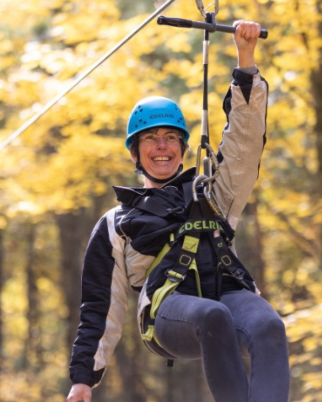 A smiling person in a blue helmet and safety harness hangs from a zip line against golden autumn trees, conveying excitement and adventure.
