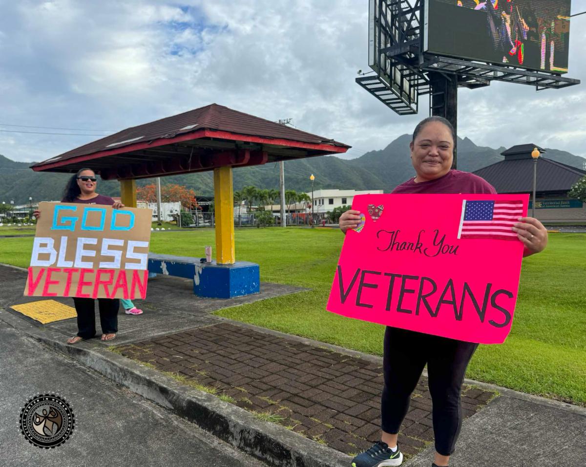 iEM students holding "Thank you Veterans" and "God Bless Veteran" signs at a Tafuna bus stop.