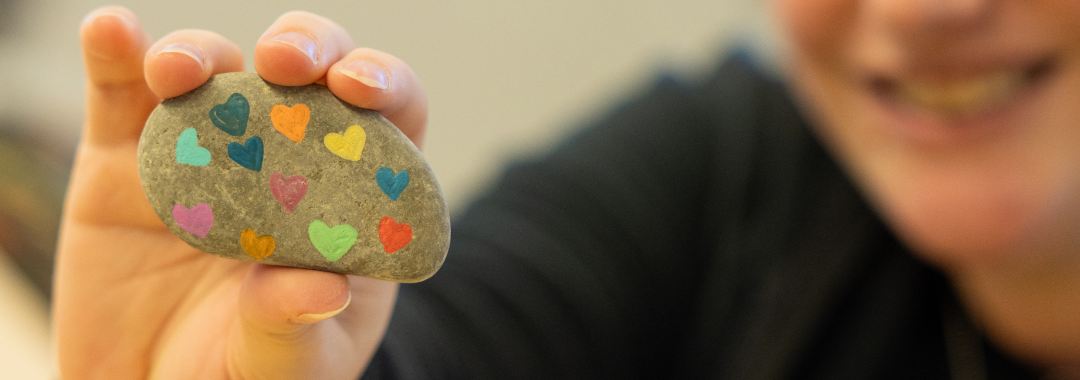 A banner image of someone holding a painted rock with hearts to the camera.