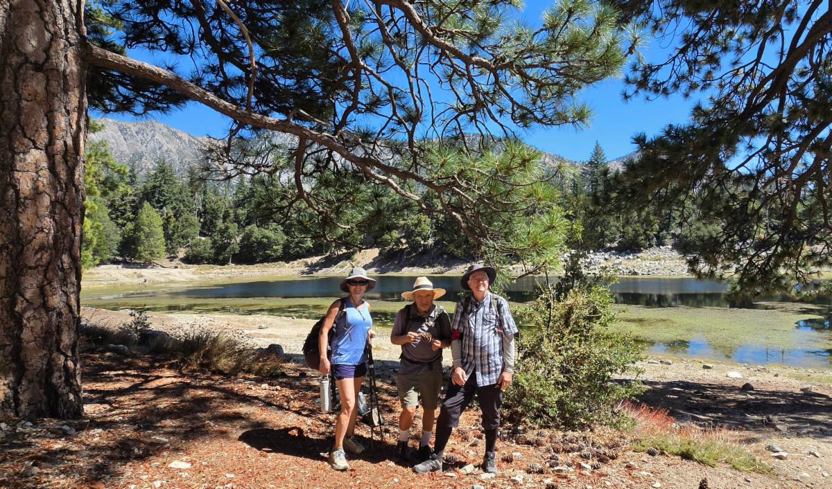 From left to right, Yvette, Hermilo, and Will wear hiking clothes and day packs on a trail beneath tall pines. Behind them, Crystal Lakeâ€™s still blue-green water reflects a wooded mountain ridge and clear blue sky.