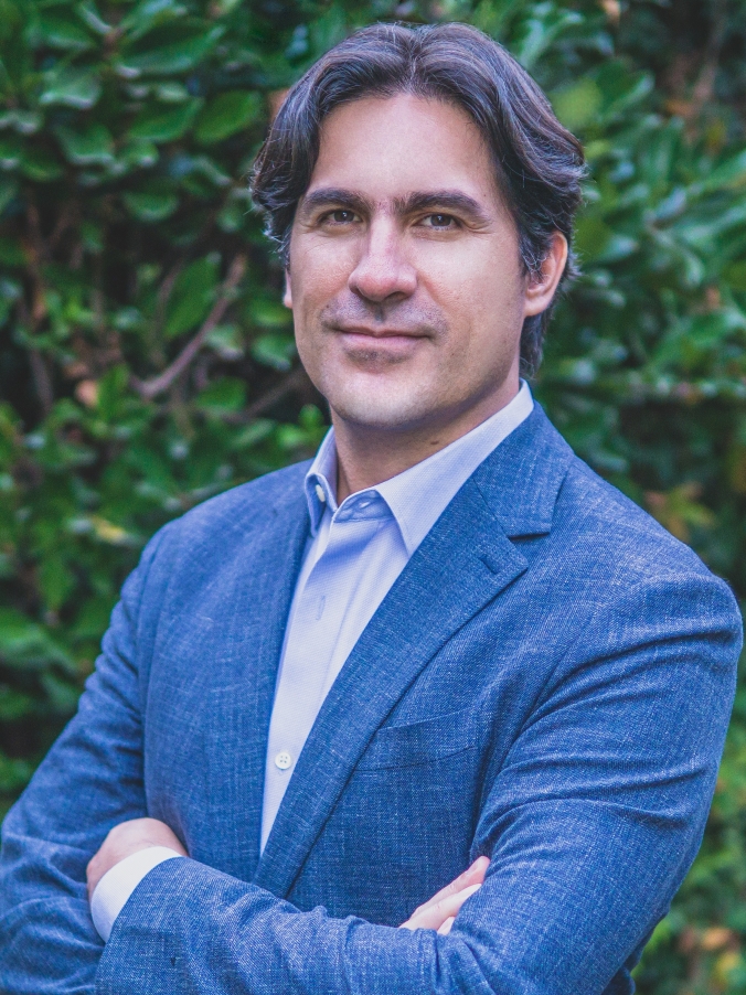 man in blue suit grinning with arms folded in front of greenery