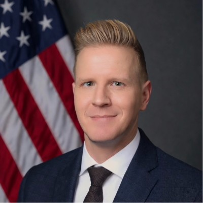 man in business suit grinning with American flag behind left shoulder