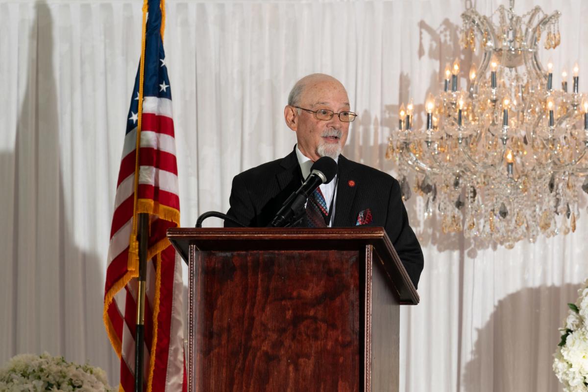 Man at podium speaking with chandelier and American flag behind him