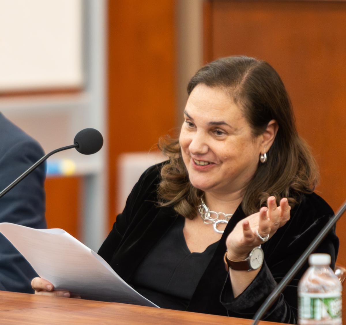 woman in black smiling and speaking into mic seated at wood table