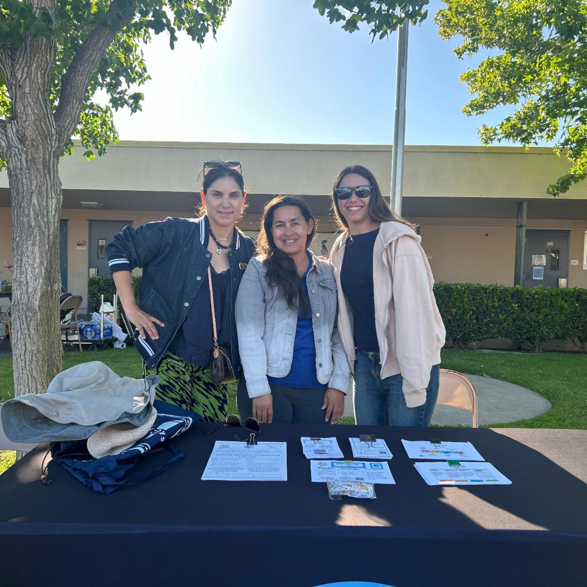 Three of our staff members standing together behind an informational table under trees