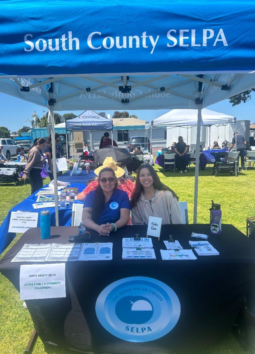 Two team members sitting behind an informational table with flyers and a tent shade