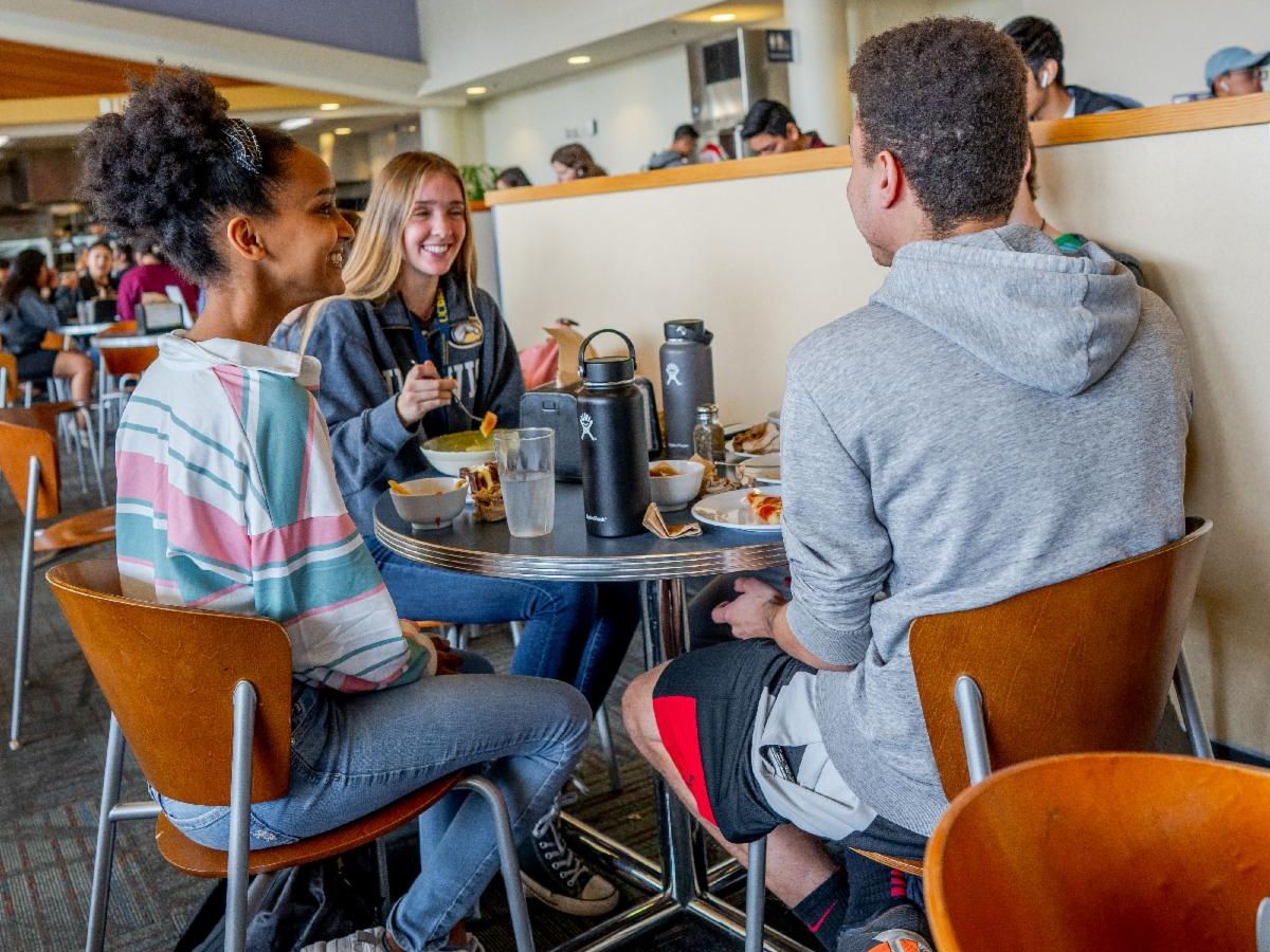 Three students sitting in the dining commons eating food.