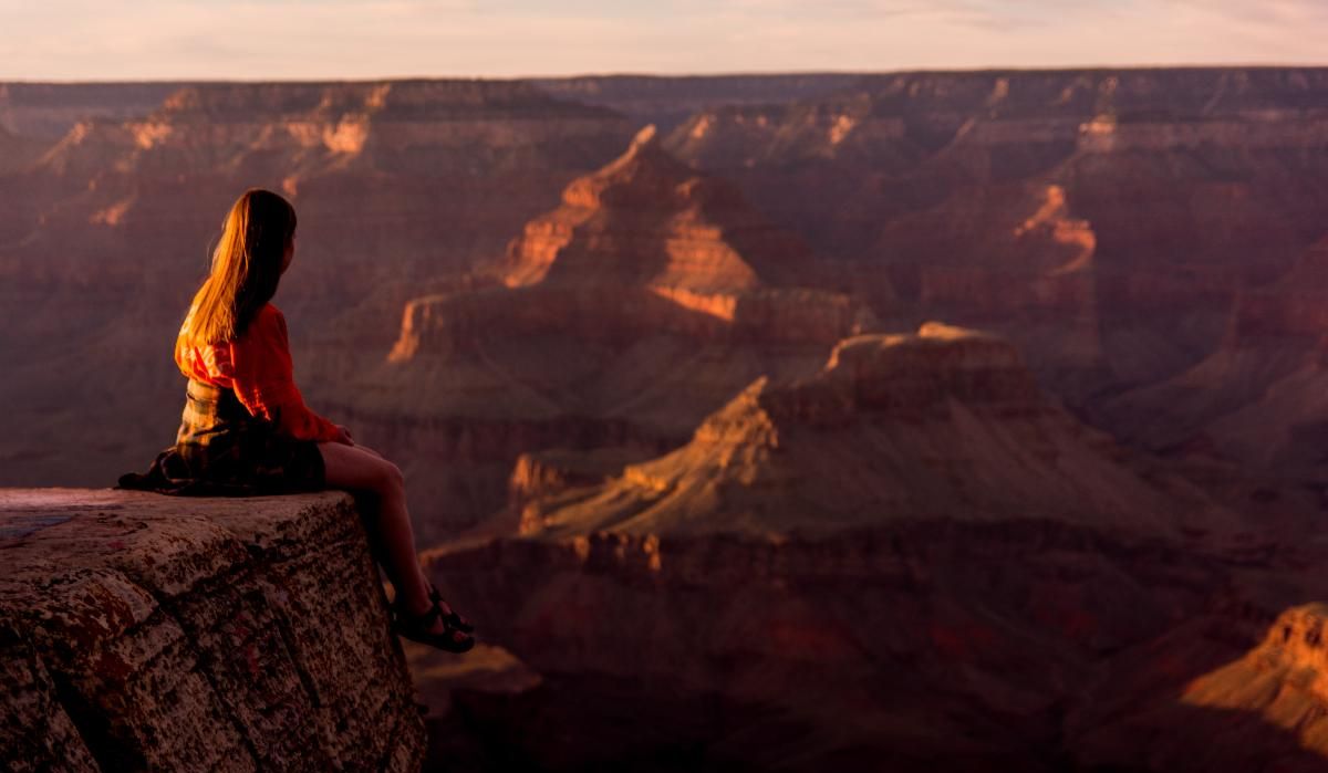 Side view of a woman sitting on the edge of the Grand Canyon as the sun sets.