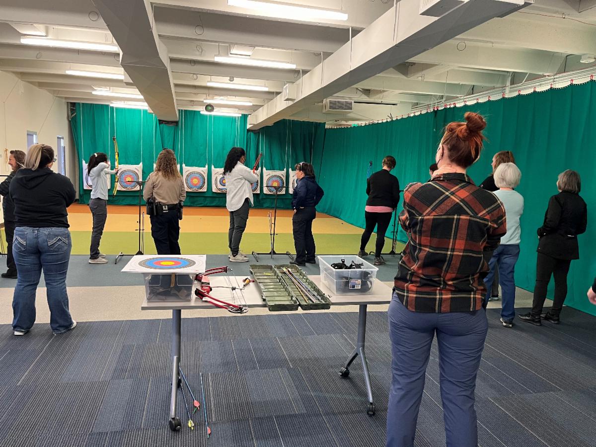 8 women lined up before their archery targets with instructors watching on.