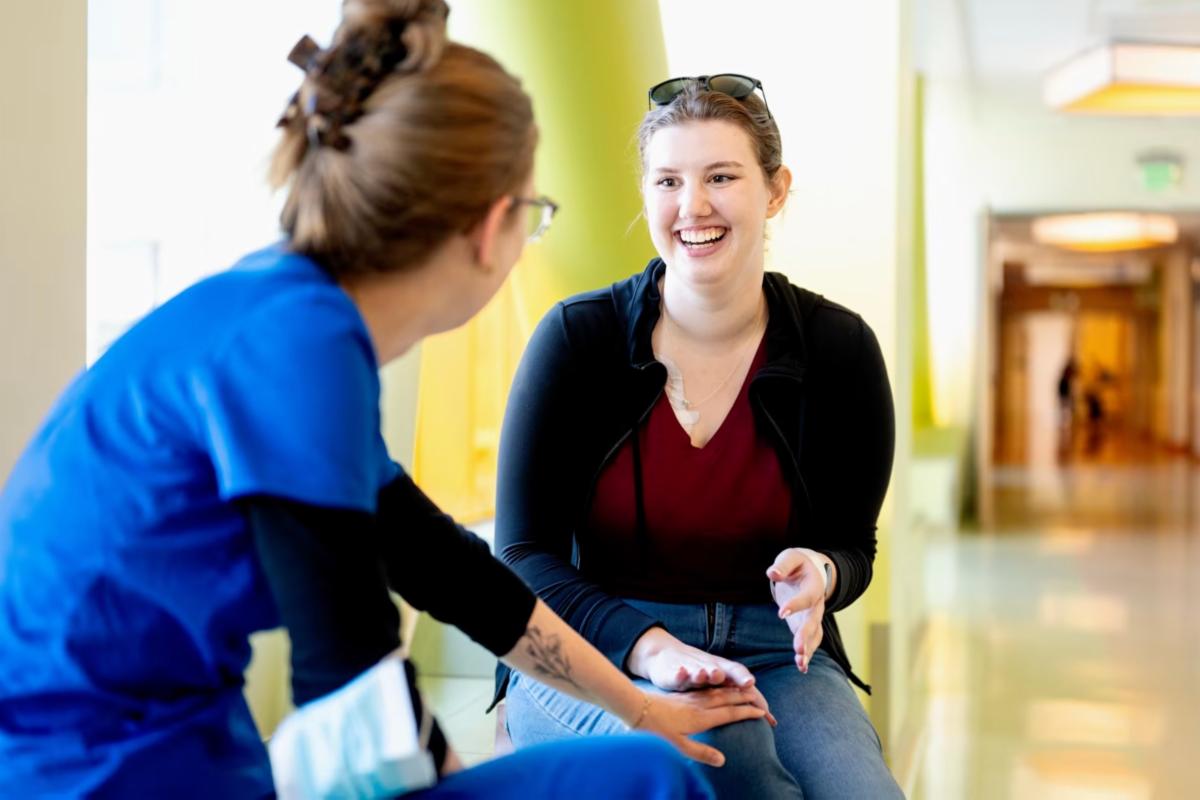 Smiling patient talks with a health care professional wearing blue scrubs in a bright hospital hallway.
