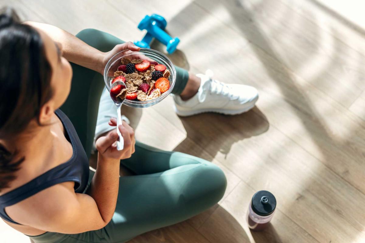 A woman in athletic gear sits while eating a bowl of fruit and oats.