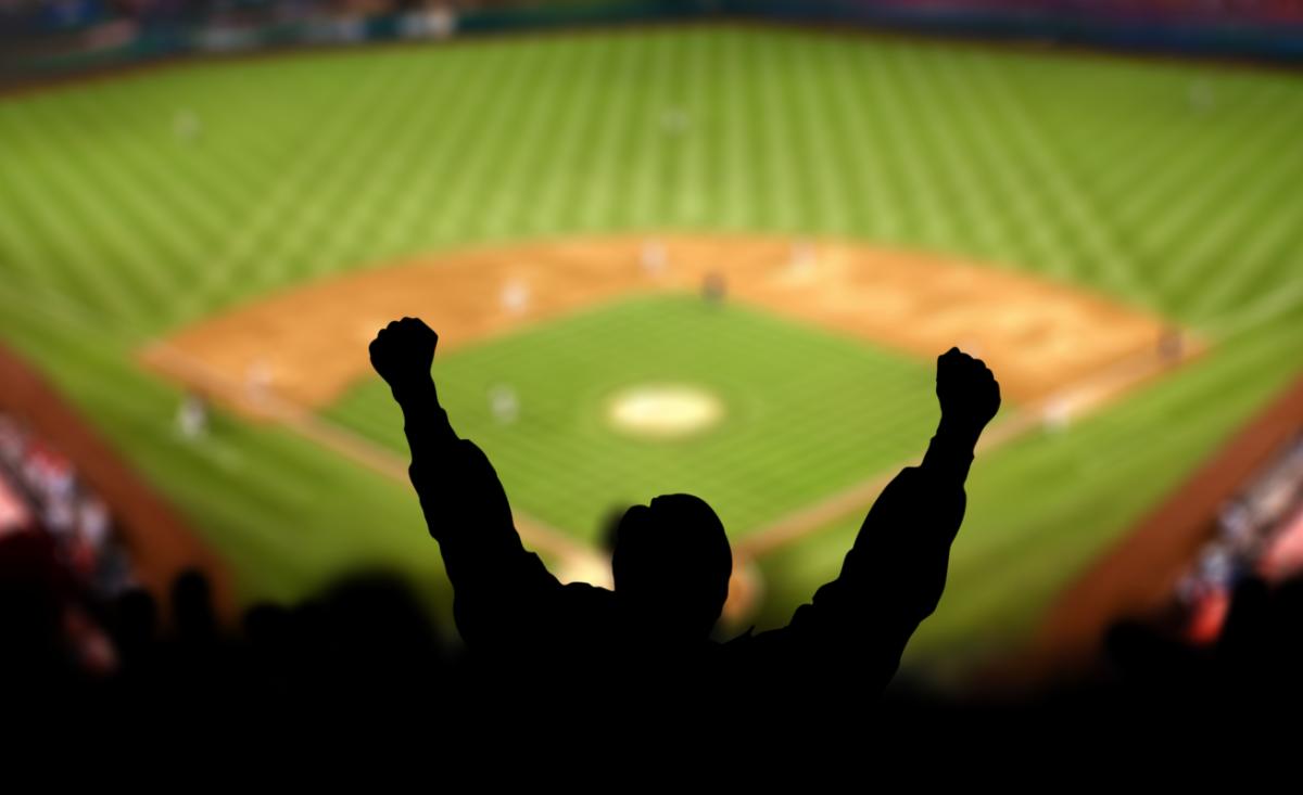 Silhouette of a baseball fan raising both arms in celebration while watching a game from the stands.