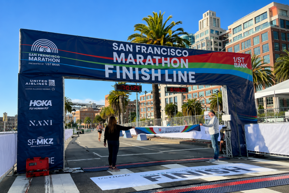 San Francisco Marathon finish line banner spanning a city street, with runners approaching the finish area and palm trees and buildings in the background.