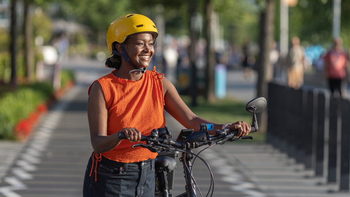 Smiling person wearing a yellow bicycle helmet stands with an e-bike on a tree-lined bike path.